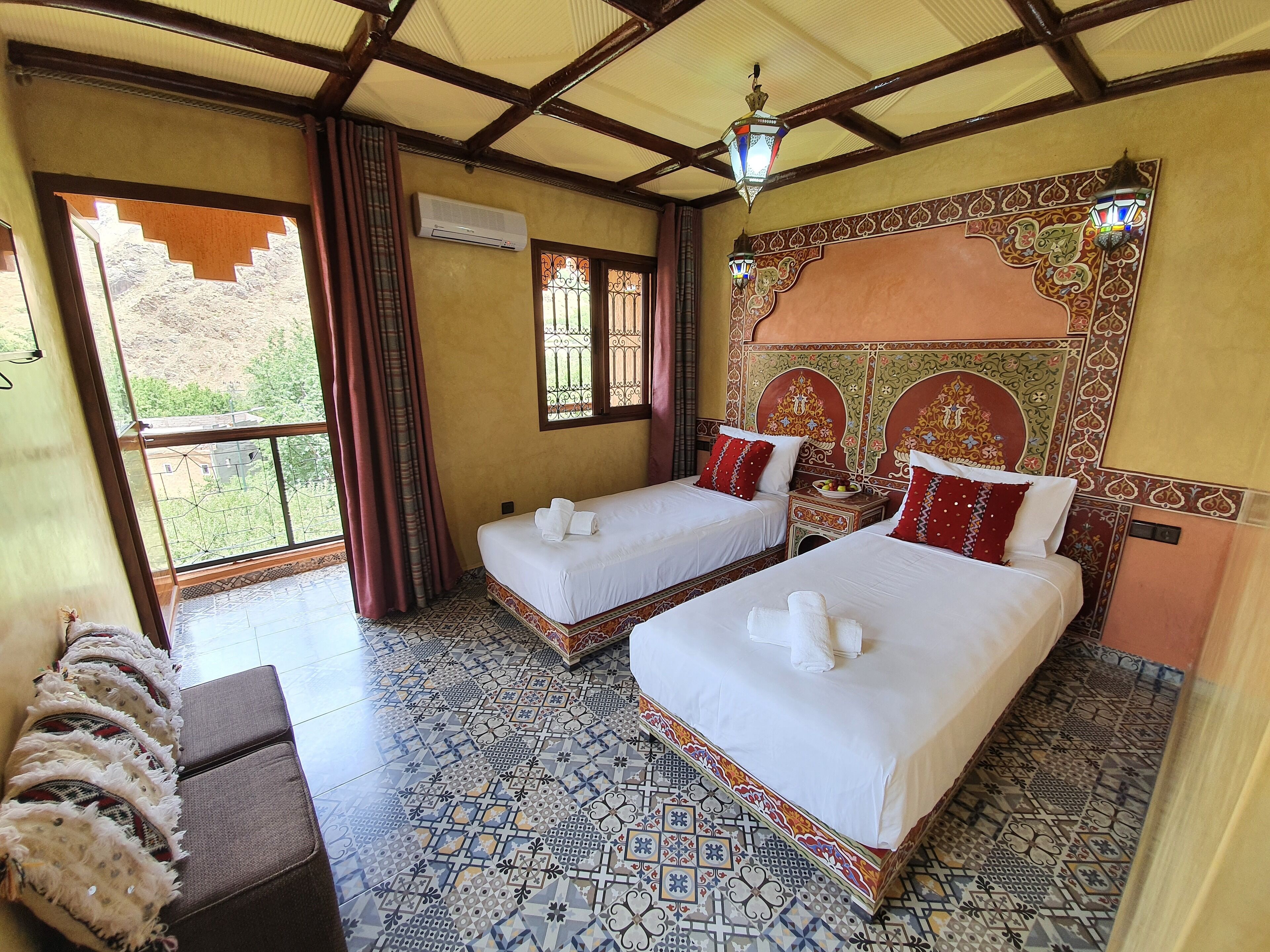 Habitación de hotel con dos camas, un ventanal con cortinas, suelo estampado y una pared decorativa.