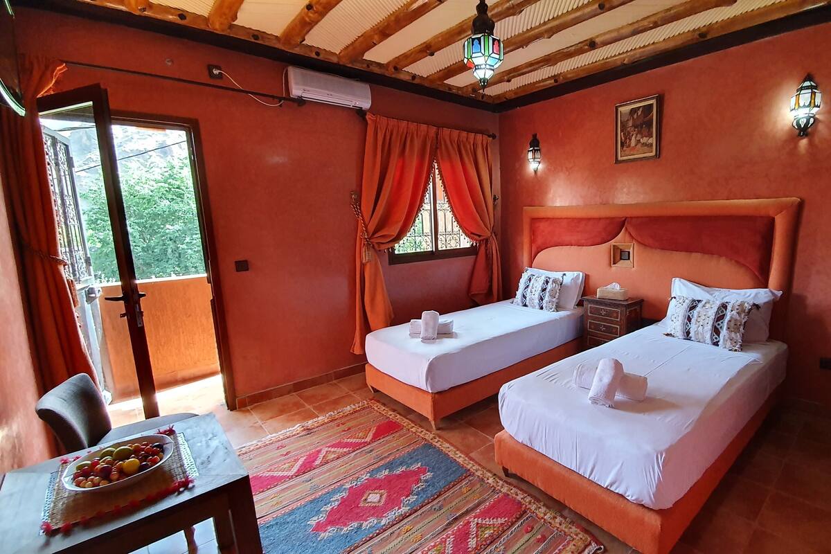 Quarto com duas camas, uma mesinha com uma fruteira, janela com cortinas e ventilador de teto.