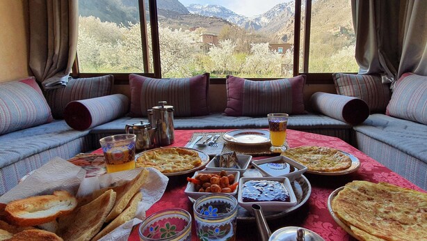 Una mesa de comedor con diversos platos y bebidas, sobre un mantel rojo. La habitación tiene un ventanal con vistas a la montaña.