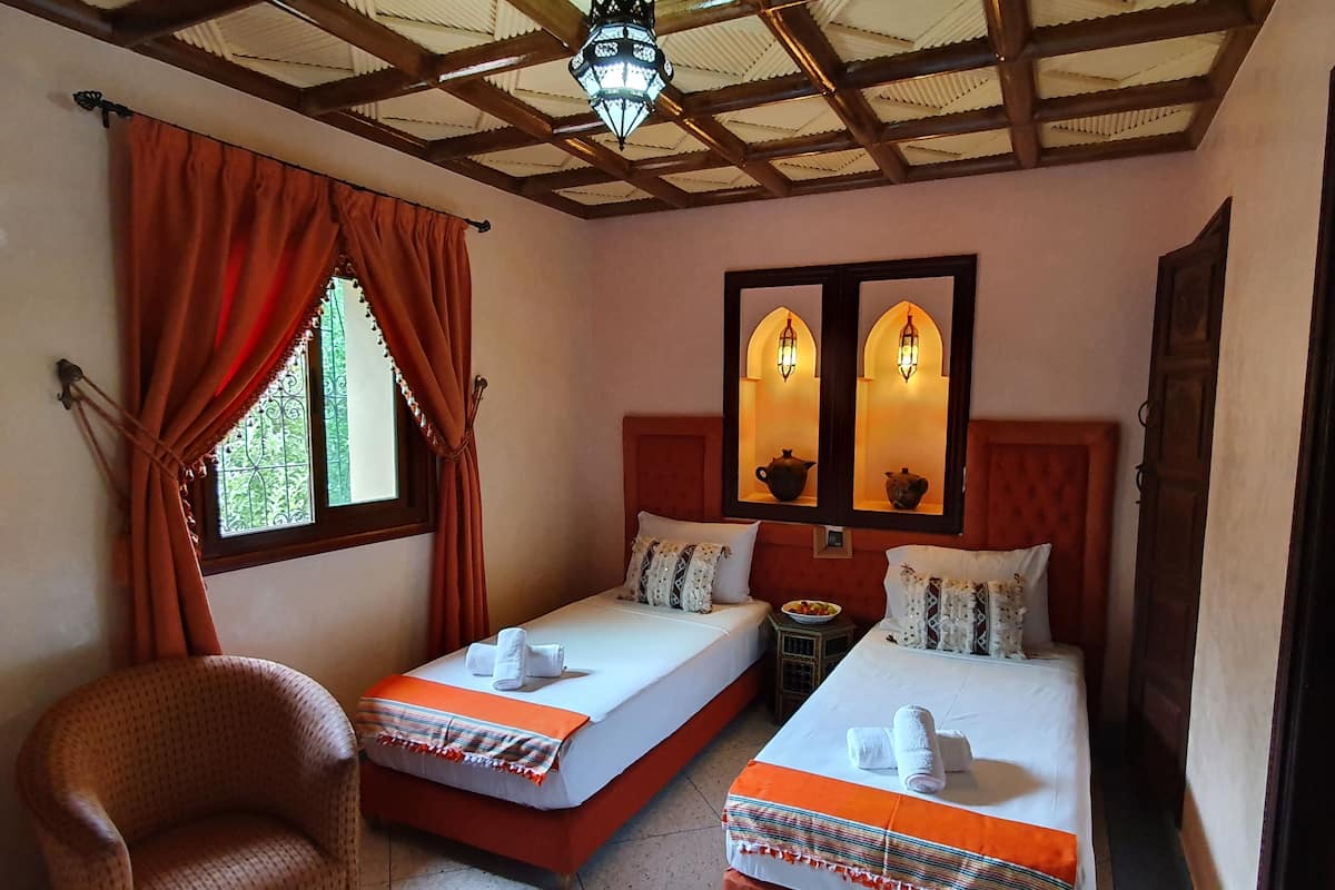 Quarto com duas camas de solteiro, uma cadeira, janela com cortinas e teto com vigas de madeira e luminária pendente.