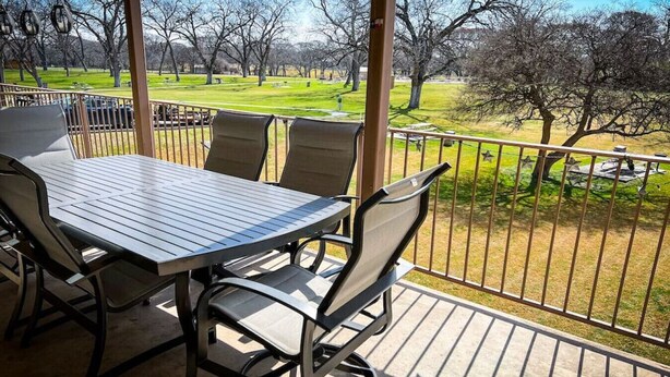 A covered outdoor patio with a table and chairs, overlooking a park with trees and grass. - Riverside Retreat | Riverfront | King Bed | Two Pools (New Braunfels)