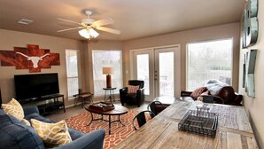 A spacious living room with a large TV, comfortable seating, a dining table, and a ceiling fan. - Riverside Retreat | Riverfront | King Bed | Two Pools (New Braunfels)