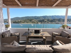 Una terraza con mobiliario de mimbre, una laptop, copas de vino y una botella, con vistas a una bahía con barcos y colinas.