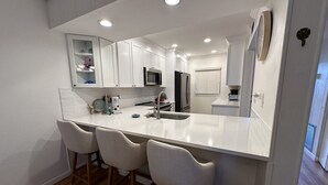 A modern kitchen with white cabinetry, a central island with bar stools, a microwave, refrigerator, and a sink. - 2 Bedroom, 2 Bathroom, Ground Floor, Pool View (Sarasota)