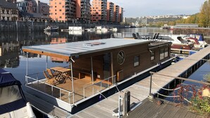 A floating houseboat docked at a marina with a view of urban buildings across the water. - Houseboat Bonanza in Prague - Bed & Cruise (Praha 7)