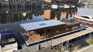 A waterfront property with a wooden deck, a small boat docked nearby, and modern buildings in the background. - Houseboat Bonanza in Prague - Bed & Cruise (Praha 7)