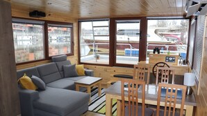 A cabin with a gray sofa, wooden dining table, and chairs, large windows with a view of boats. - Houseboat Bonanza in Prague - Bed & Cruise (Praha 7)
