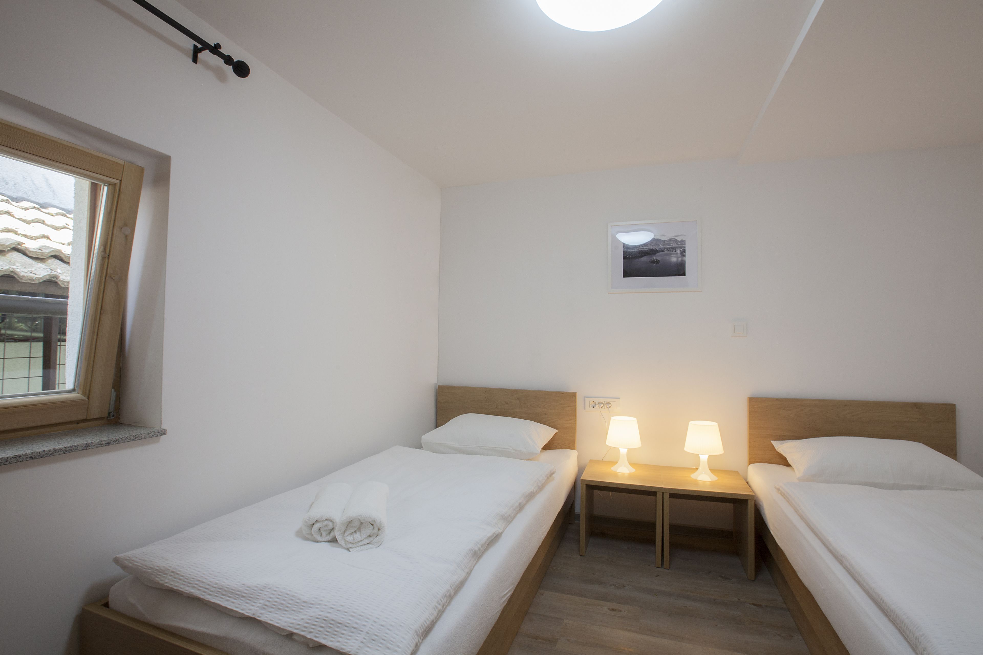 Quarto com duas camas de solteiro, mesa de cabeceira com luminária, pequena imagem na parede e janela com moldura de madeira.