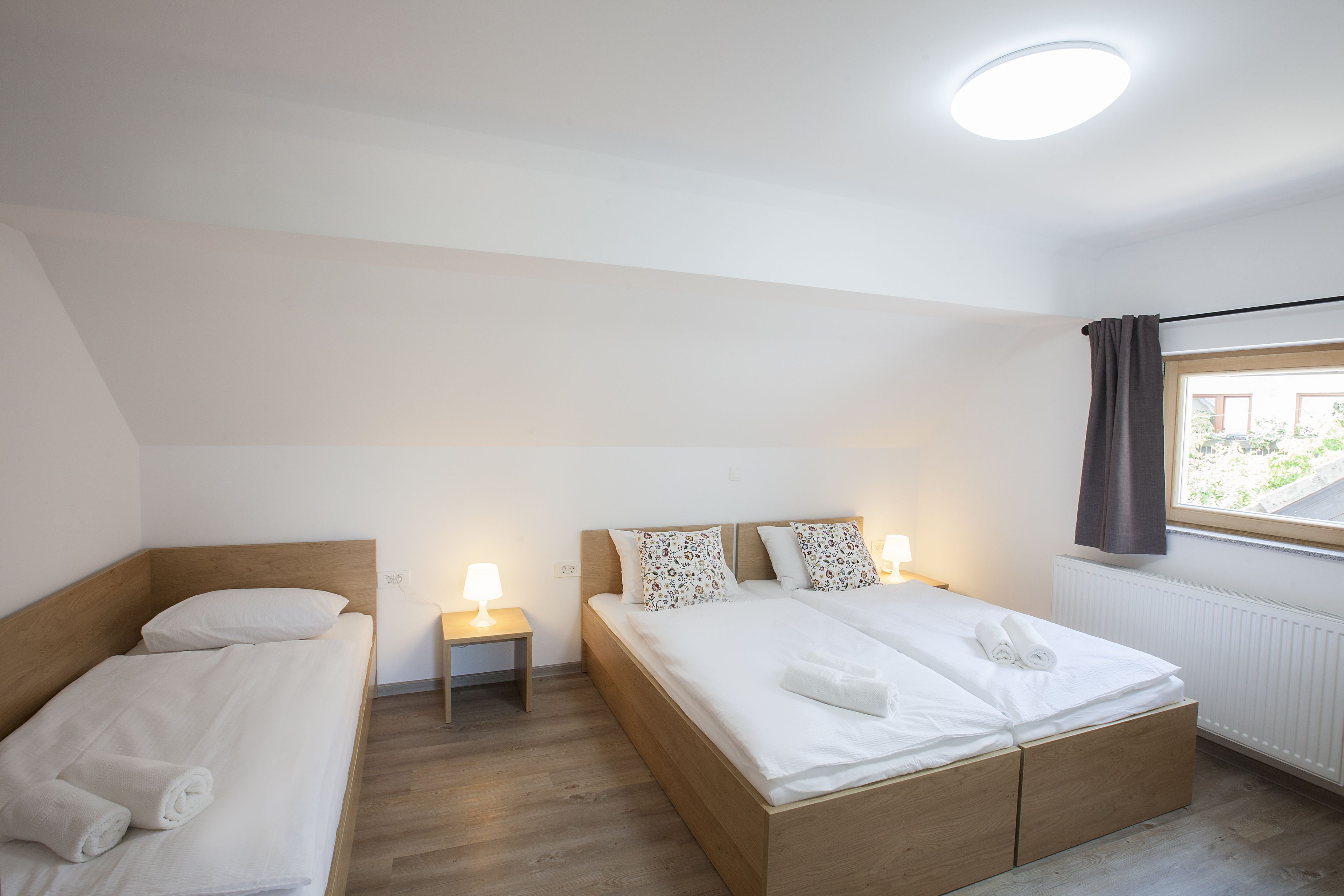 A hotel room with two single beds, a large bed, a window with curtains, and a nightstand with a lamp.