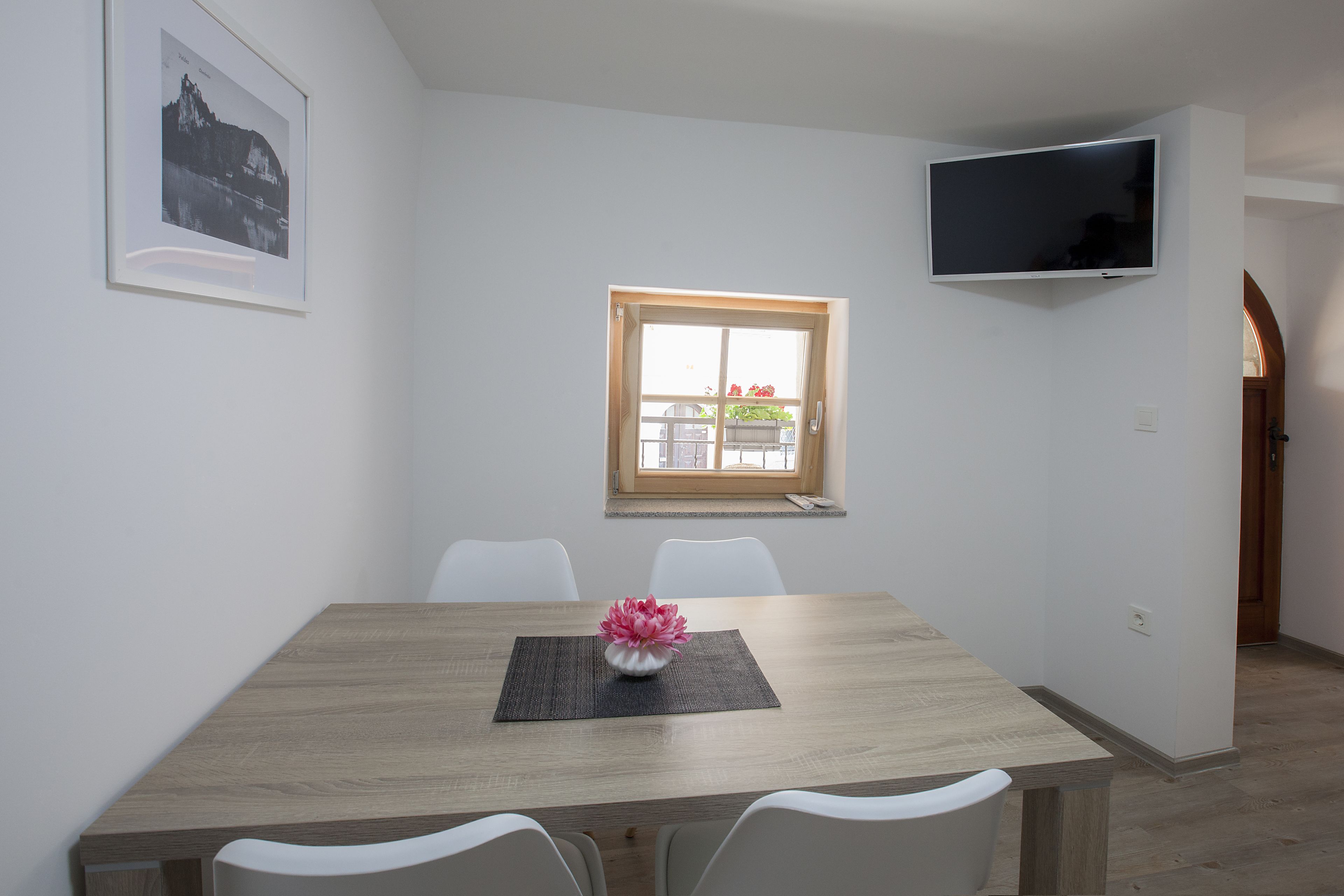 A dining area with a wooden table, white chairs, a flower vase, a framed picture, a window, and a TV mounted on the wall.