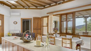 A spacious kitchen with wooden beams, a large island with a cheese and wine, and a view through the windows. - Naxos Cottage - Cottage 1 (Naxos)
