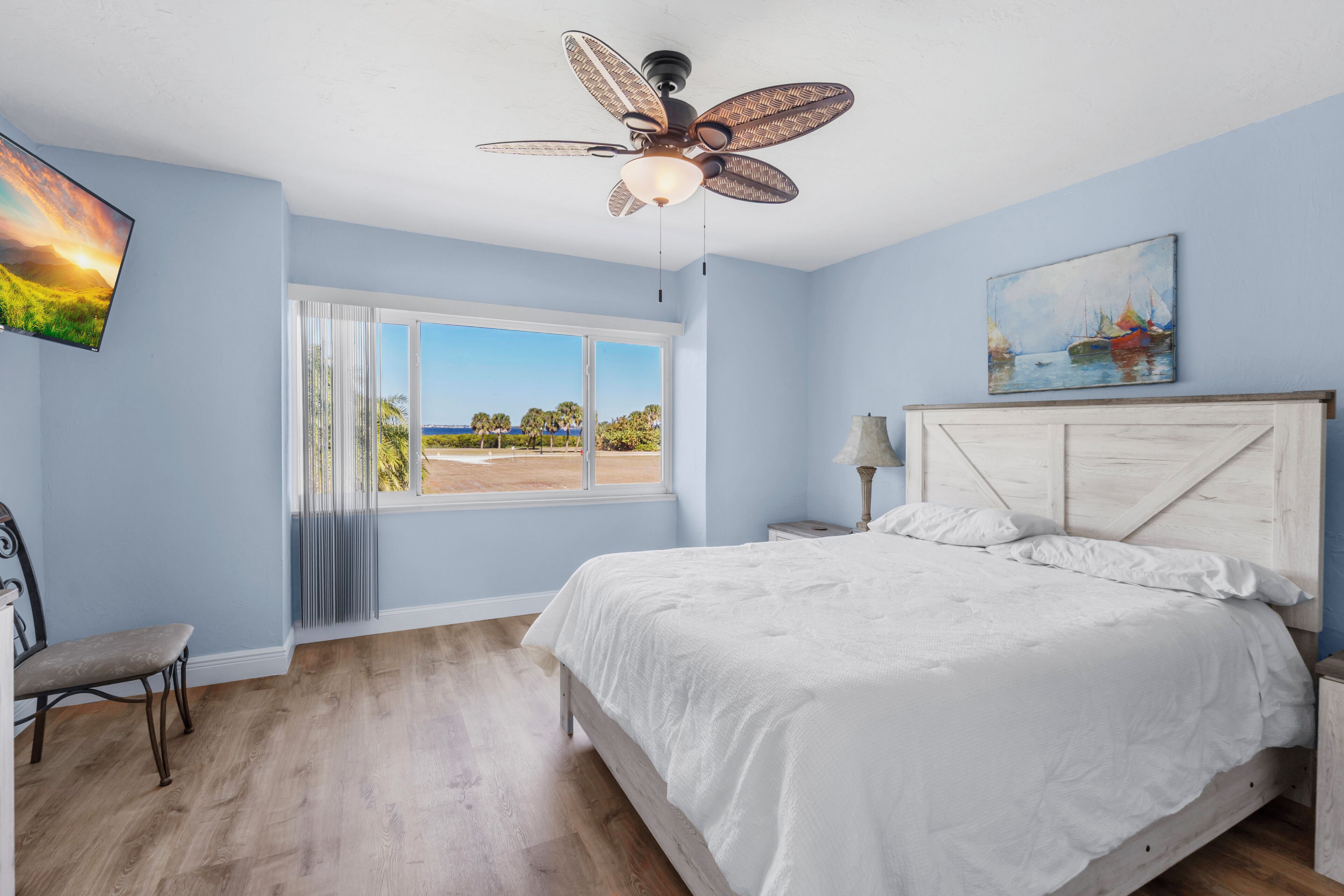 A bedroom with a bed, a ceiling fan, a window with a view, and a painting on the wall.