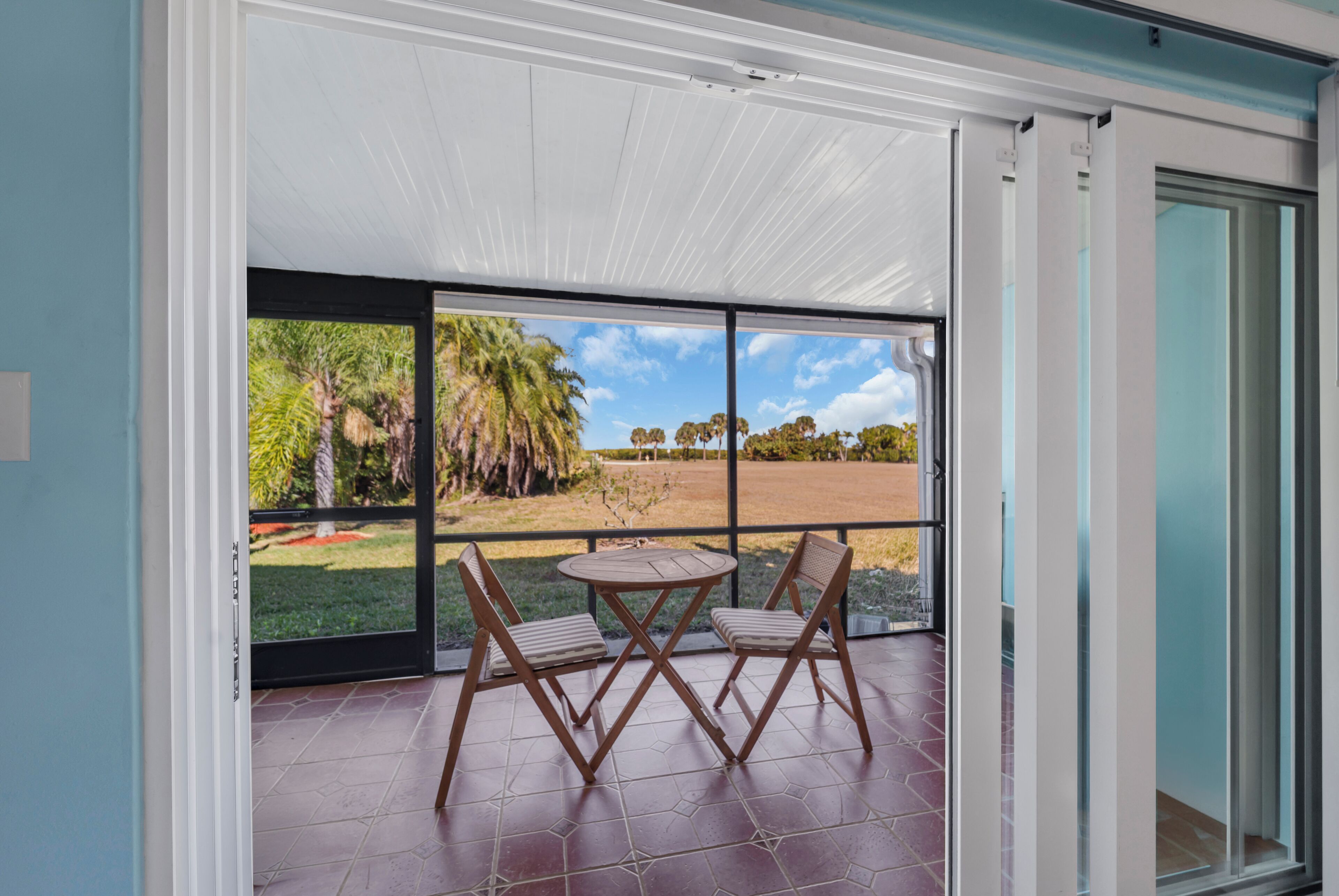 Une pièce dotée d’une porte-fenêtre coulissante donnant sur un patio avec une table et des chaises, un sol carrelé et une vue sur un espace verdoyant avec des arbres.