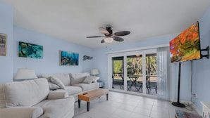Una sala de estar con sofá, una mesa de centro, un ventilador de techo y un ventanal con vistas al exterior.