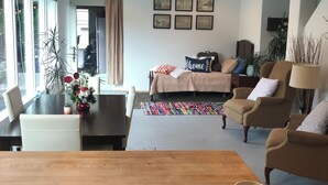 Ein modernes Wohnzimmer mit Couch, Sesseln und einem Holztisch. An den Wänden hängen Bilderrahmen, und es gibt einen Esstisch mit Stühlen sowie ein großes Fenster mit Vorhängen.