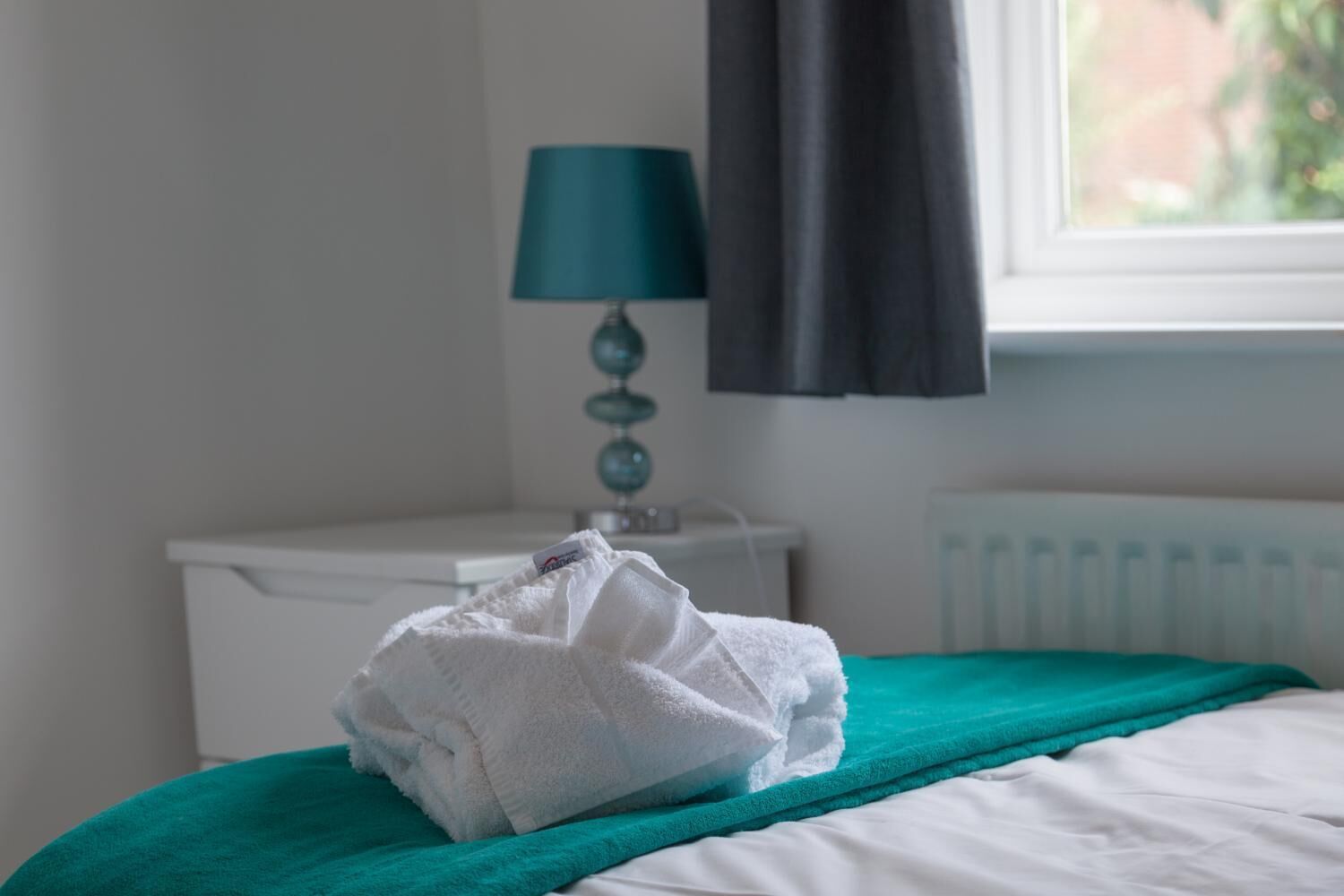 A neatly made bed with a teal blanket and a folded white towel on it.