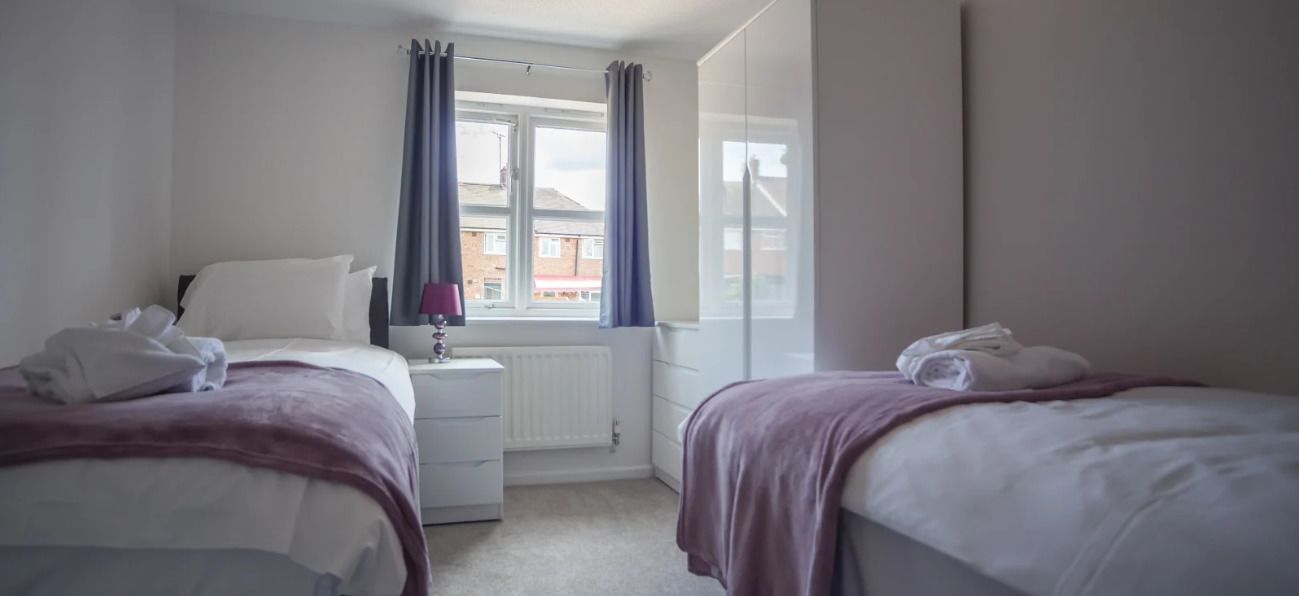 A hotel room with two beds, a window with curtains, a radiator, and a view of buildings outside.
