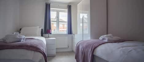 A hotel room with two beds, a window with curtains, a radiator, and a view of buildings outside.
