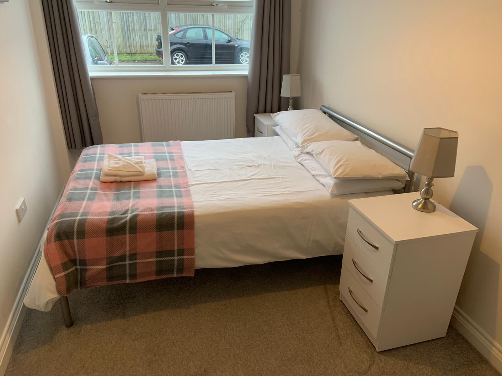 A neatly made bed with a plaid blanket and a folded towel, a bedside lamp, and a nightstand with drawers.