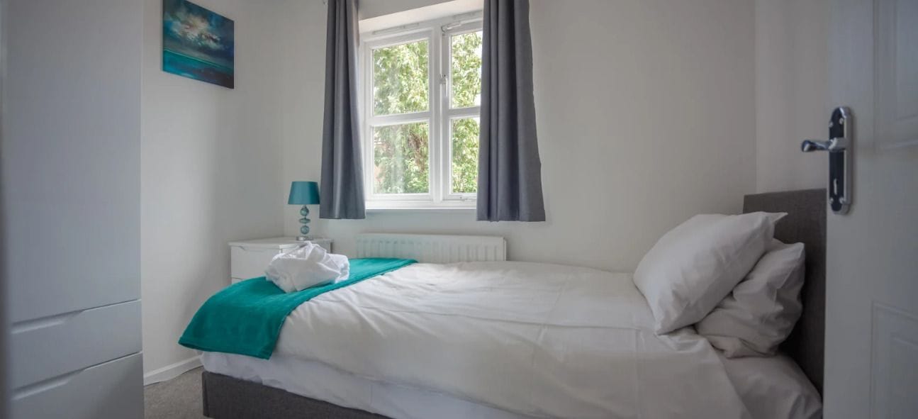 A bedroom with a bed, a nightstand, a window with curtains, a radiator, and a door.