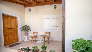 Eine Holztür, eine weiße Wand mit einem gerahmten Bild, zwei Holzstühle, ein kleiner weißer Tisch und mehrere Blumentöpfe.