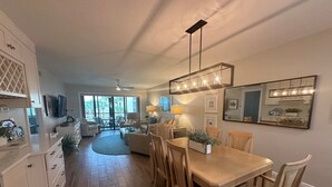 A spacious living area with a wooden dining table, comfortable seating, and a large window offering a view of the outdoors. - Renovated, Gulf View Unit, Overlooking the Pool and Beautiful Courtyard (Sarasota)