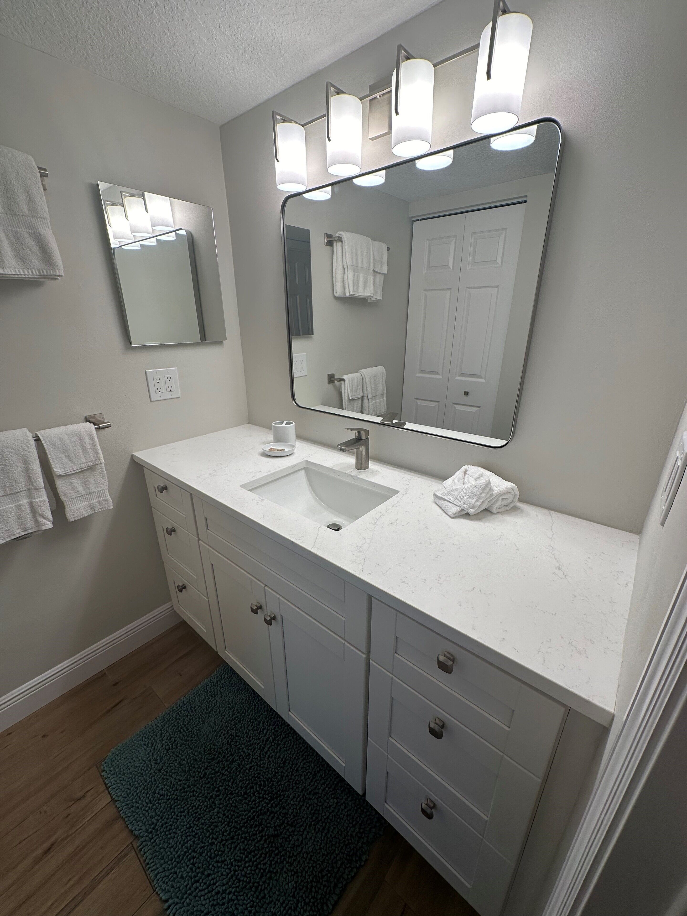 A bathroom with a white sink, a large mirror, and a vanity with drawers. There are towels hanging on the wall and a green rug on the floor.
