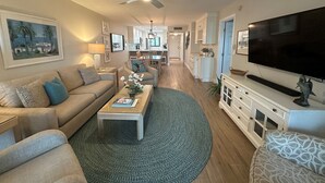 A spacious living room with a sofa, coffee table, and TV. There is a kitchen in the background and a hallway leading to other rooms. - Renovated, Gulf View Unit, Overlooking the Pool and Beautiful Courtyard (Sarasota)