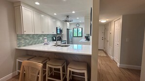 A modern kitchen with white cabinets, a large island with stools, stainless steel appliances, and a clock on the wall. - Renovated, Gulf View Unit, Overlooking the Pool and Beautiful Courtyard (Sarasota)