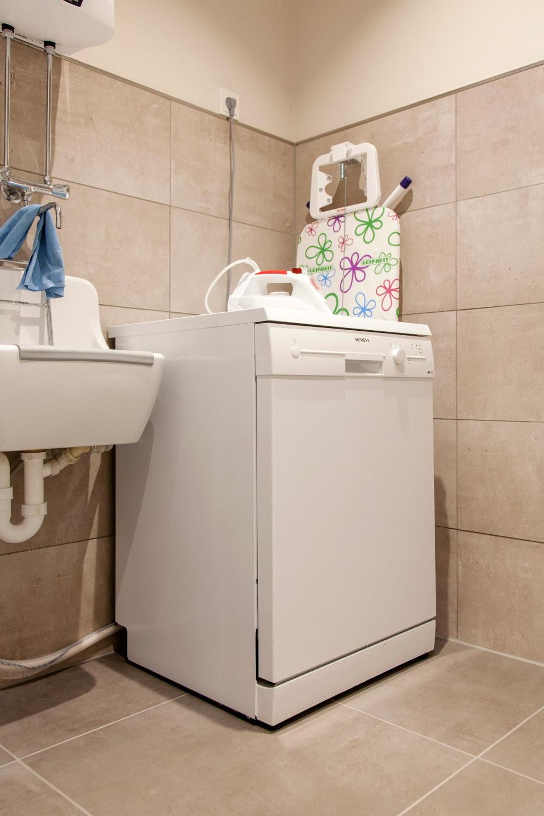 Une machine à laver blanche et compacte, dotée d’un panneau de commande et d’un couvercle coloré, est placée à côté d’un évier dans une salle de bain aux carreaux beiges, où une serviette bleue est suspendue au robinet.