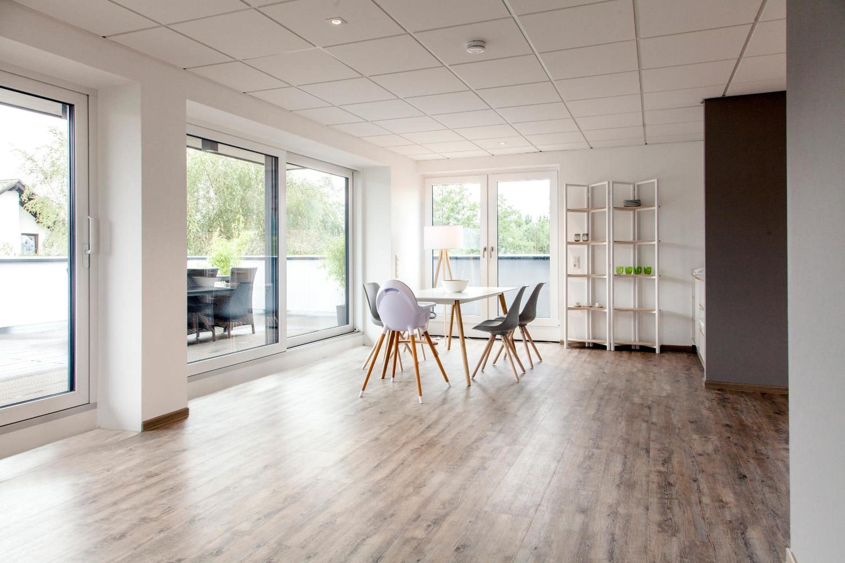 A spacious room with a large window, a dining area with chairs and a table, and a white shelving unit.