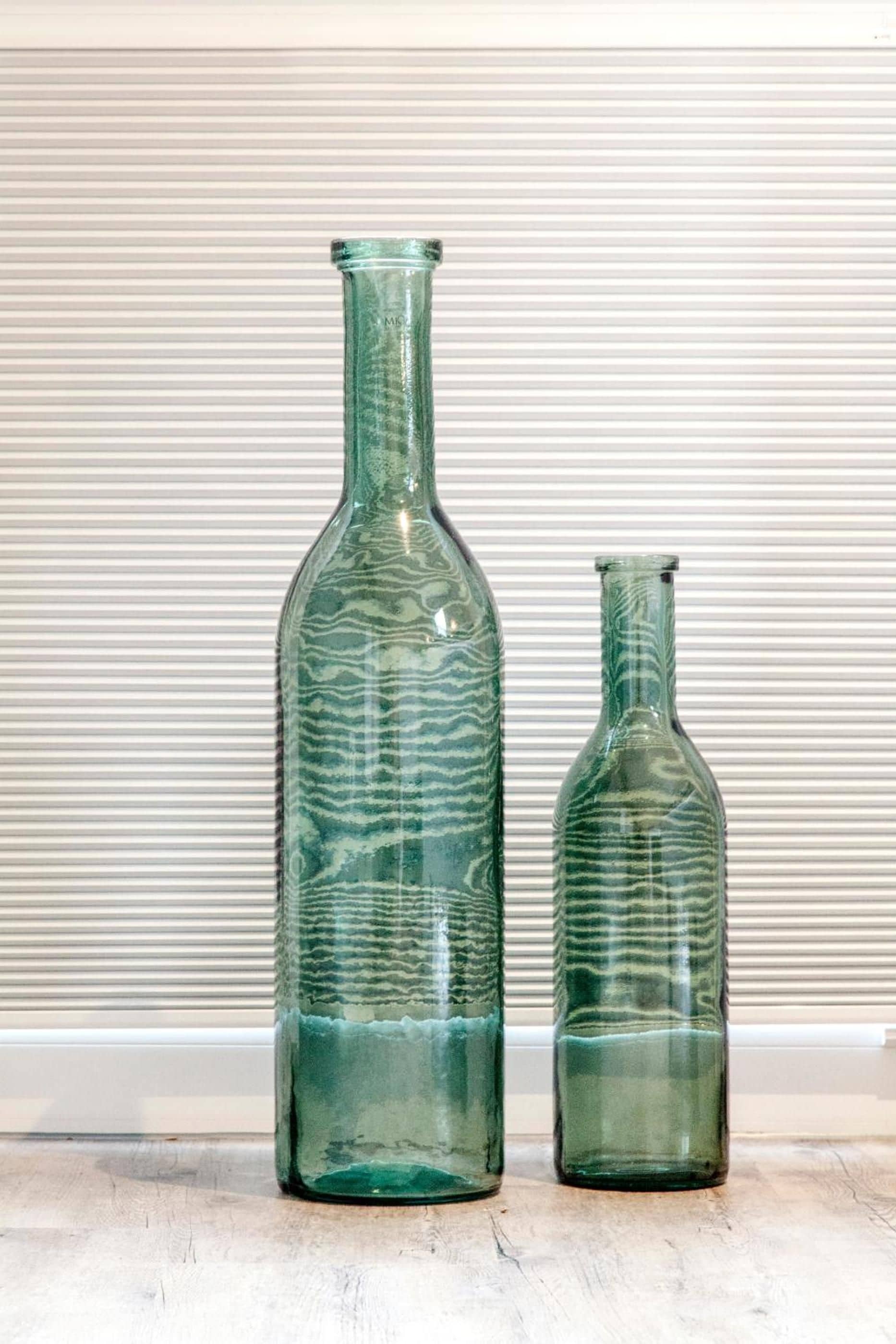 Deux bouteilles en verre, d’une teinte verdâtre et dotées d’un motif texturé, sont posées sur une surface en bois, le tout sur un fond à rayures.