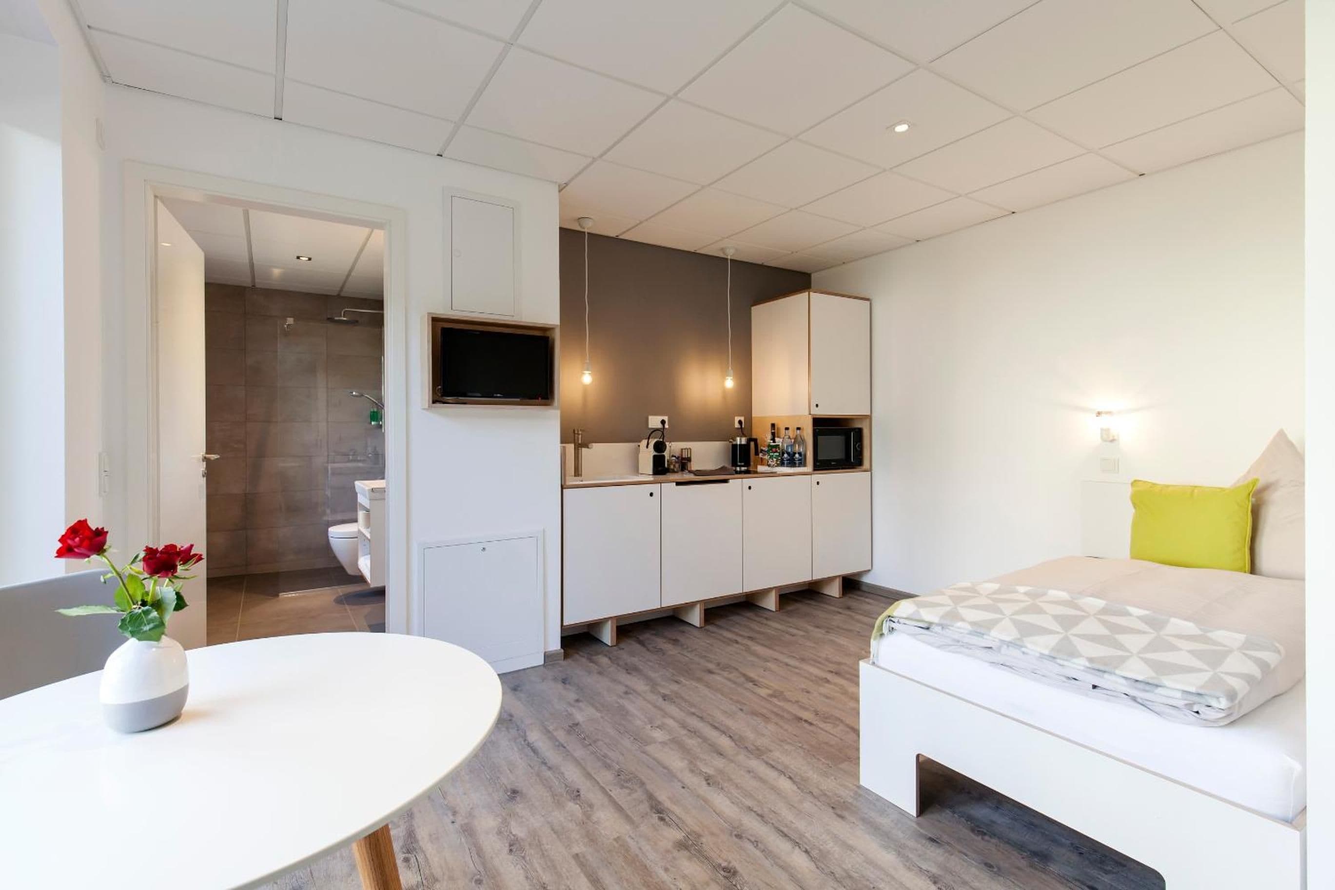 Una habitación de hotel moderna con cama, una kitchenette pequeña, baño y área de comedor.