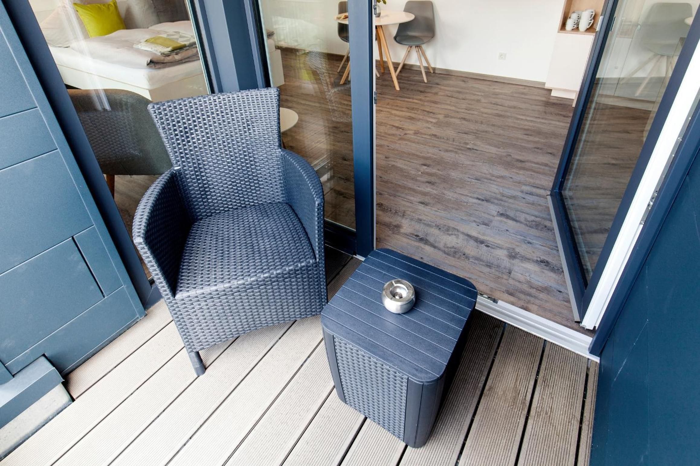 A balcony with a wicker chair and a small table, wooden flooring, and a glass door leading to an indoor area.