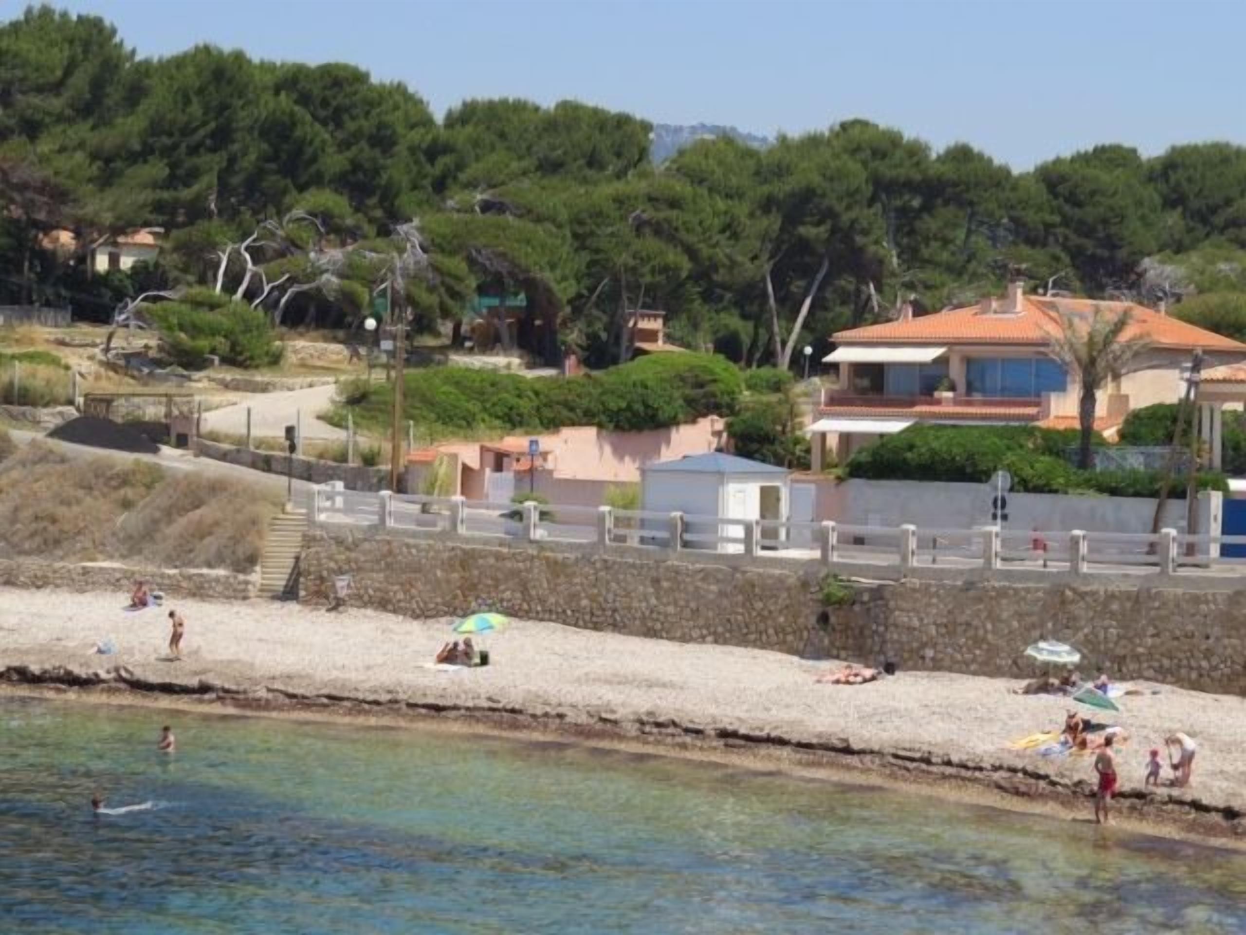 Ett kustområde med en sandstrand, en stenmur och hus i bakgrunden.