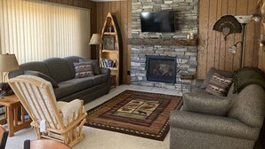 A cozy living room with a stone fireplace, a flat-screen TV mounted above it, and a comfortable seating area with two sofas and a wooden chair.