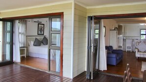 A room with a bed, a small table, and a window with curtains. There is a door leading to another room with a sofa and a table. - Alfies House (Ponta d'Ouro)