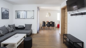 Un salon moderne avec un canapé gris, une table basse blanche et un banc noir. On aperçoit à l’arrière-plan un coin repas avec une table et des chaises en bois.
