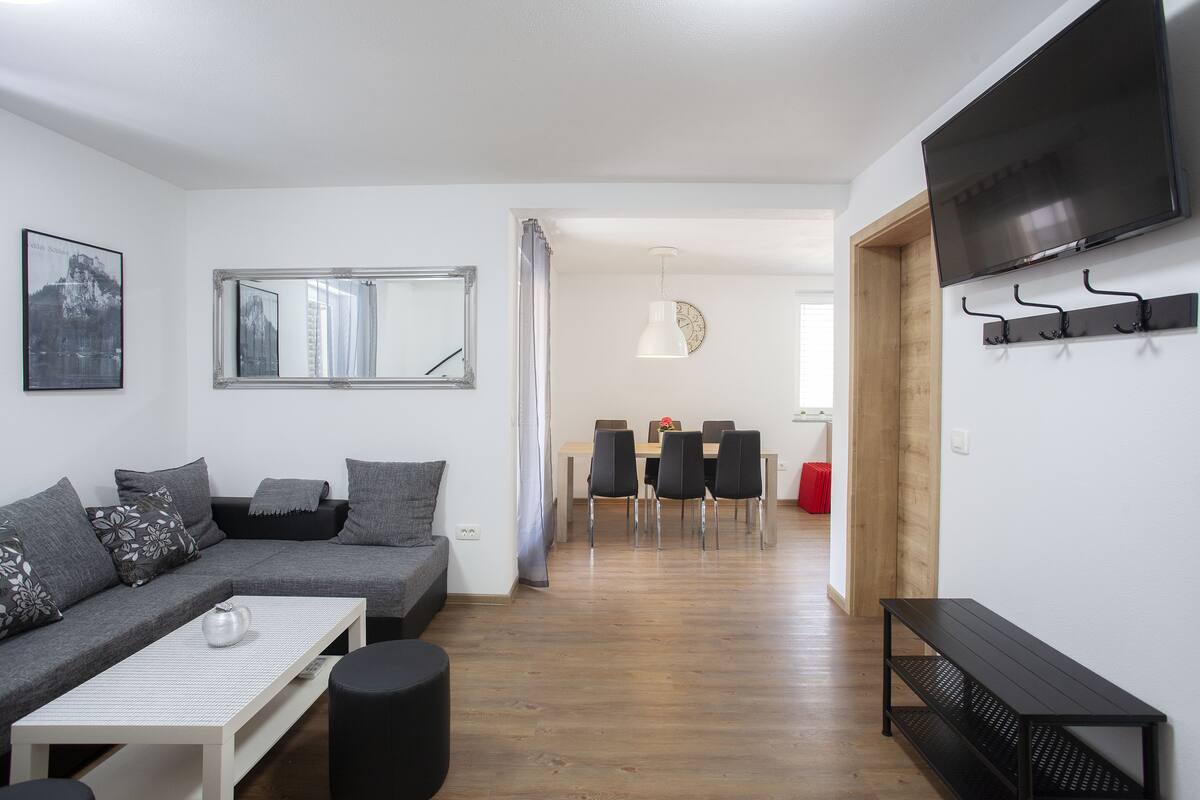 Una sala moderna con un sofá gris, una mesa de centro blanca y un banco negro. En el fondo, se observa un comedor con una mesa y sillas de madera.