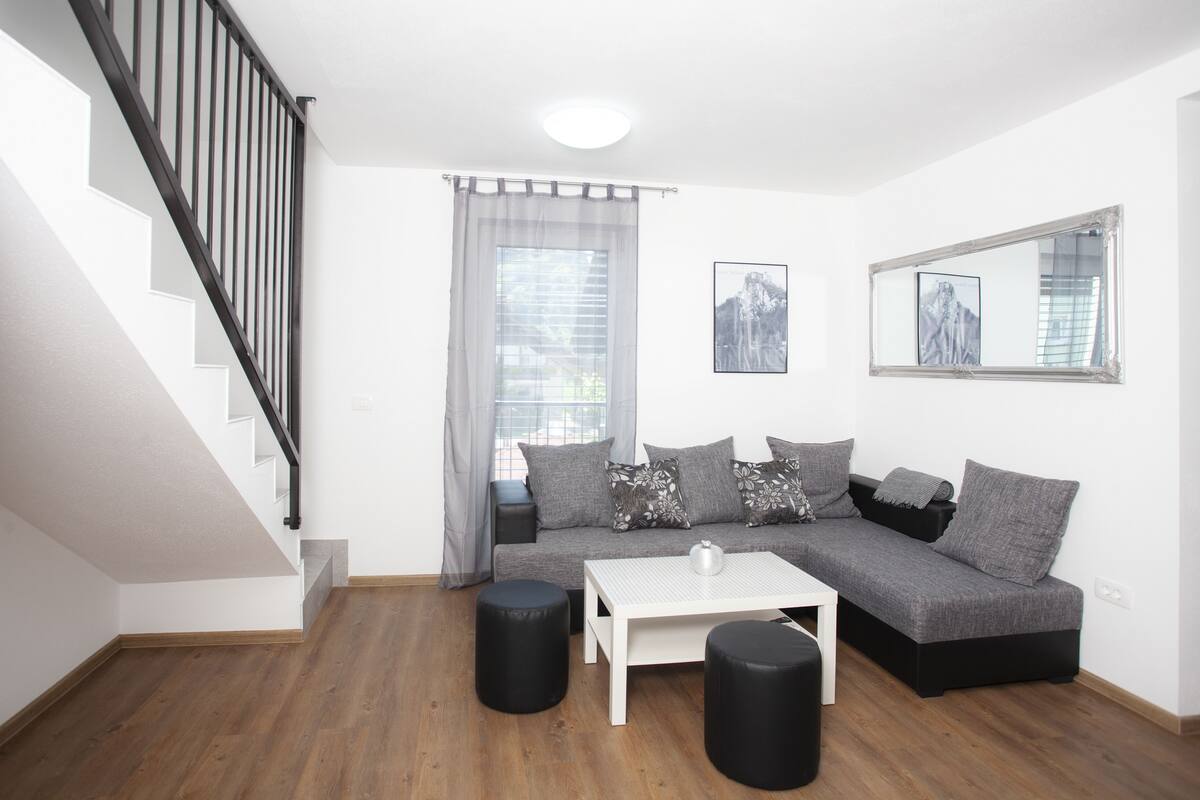 Una sala de estar moderna con una escalera, un sofá gris, otomanas negras y una mesa de centro blanca.