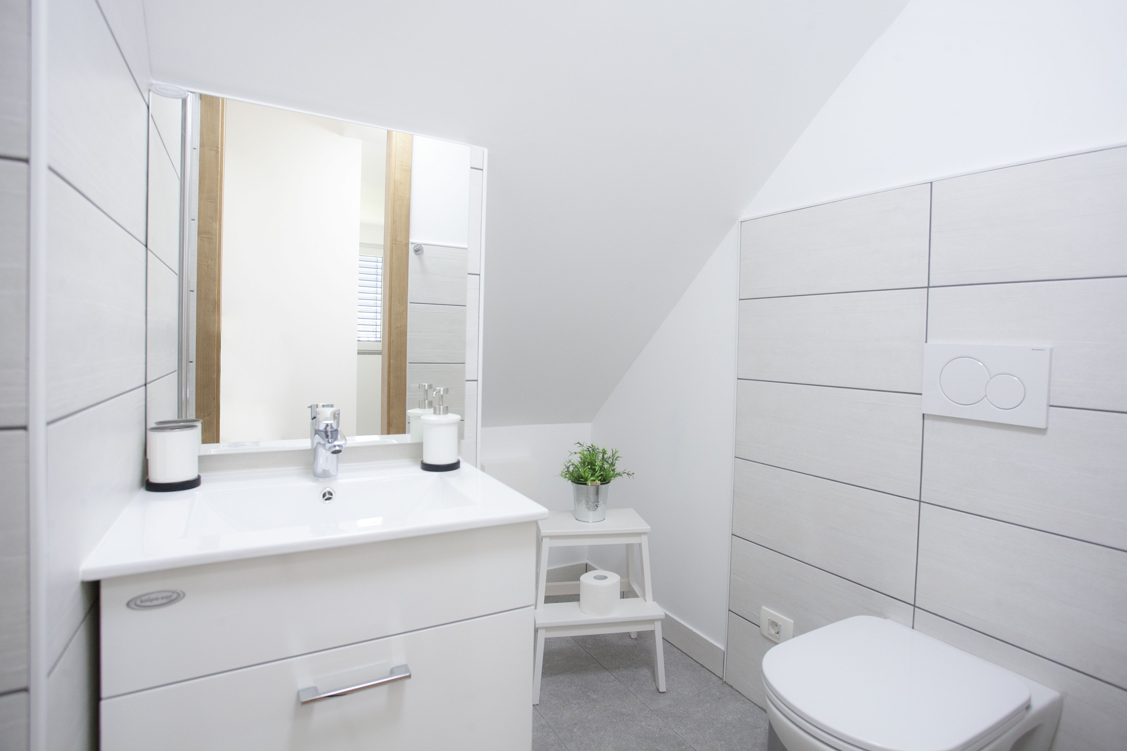 A modern bathroom with a white vanity, a mirror, a toilet, and a small step stool with a plant.