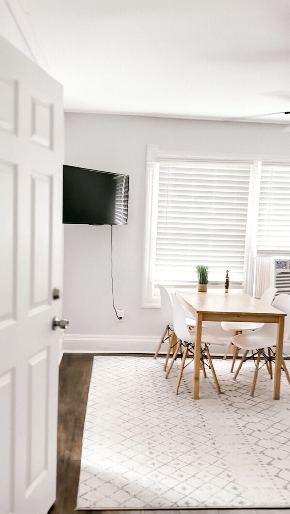 A dining area with a table and chairs, a mounted television, and a window with blinds.