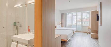 A hotel room with a glass shower, a white sink, two beds, a wooden desk, and a large window with a view of buildings.