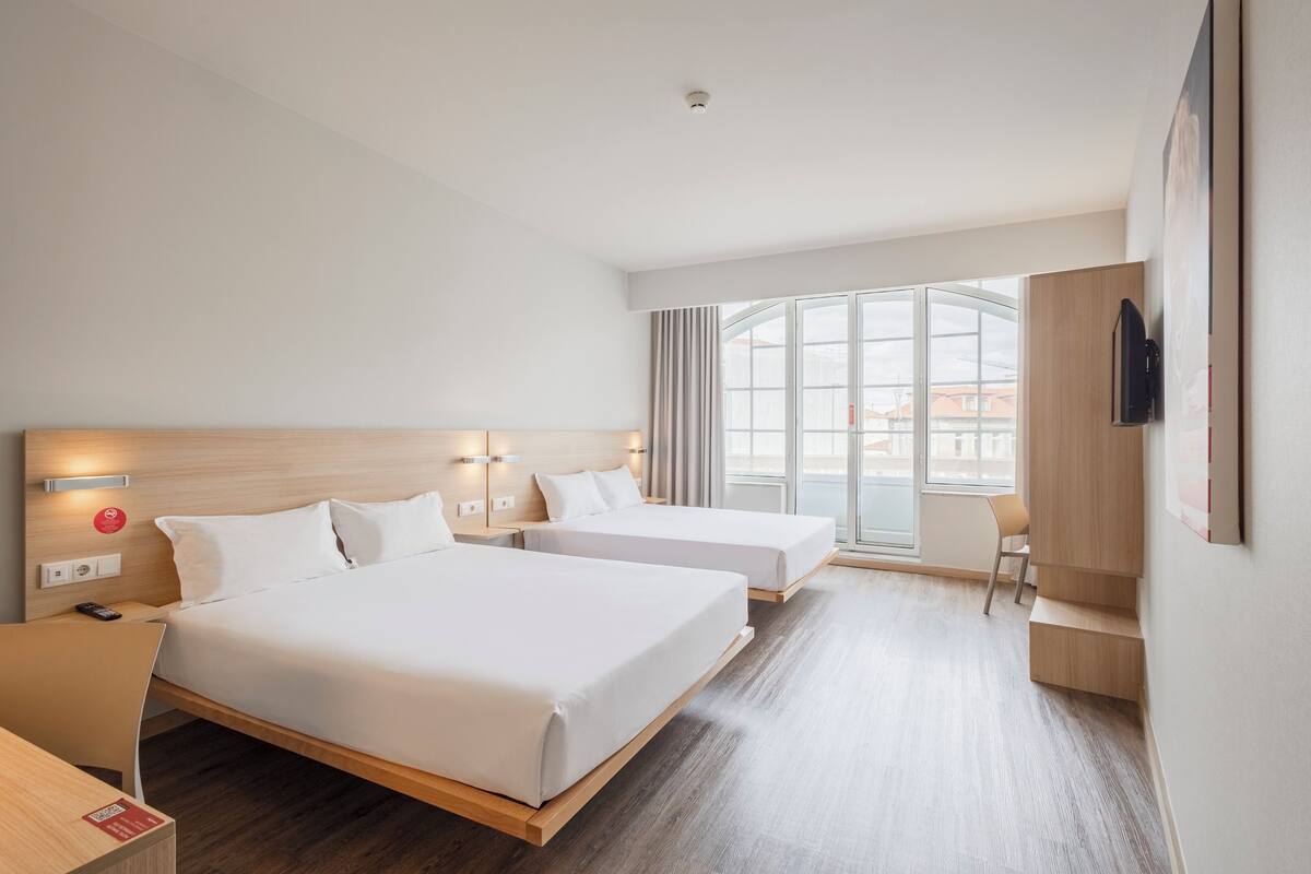 A hotel room with two beds, a desk, a chair, a TV, and a large window.