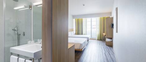 Habitació d'hotel moderna amb dutxa de vidre, un lavabo blanc, dos llits, un armari de fusta i una finestra gran amb cortines a ratlles.