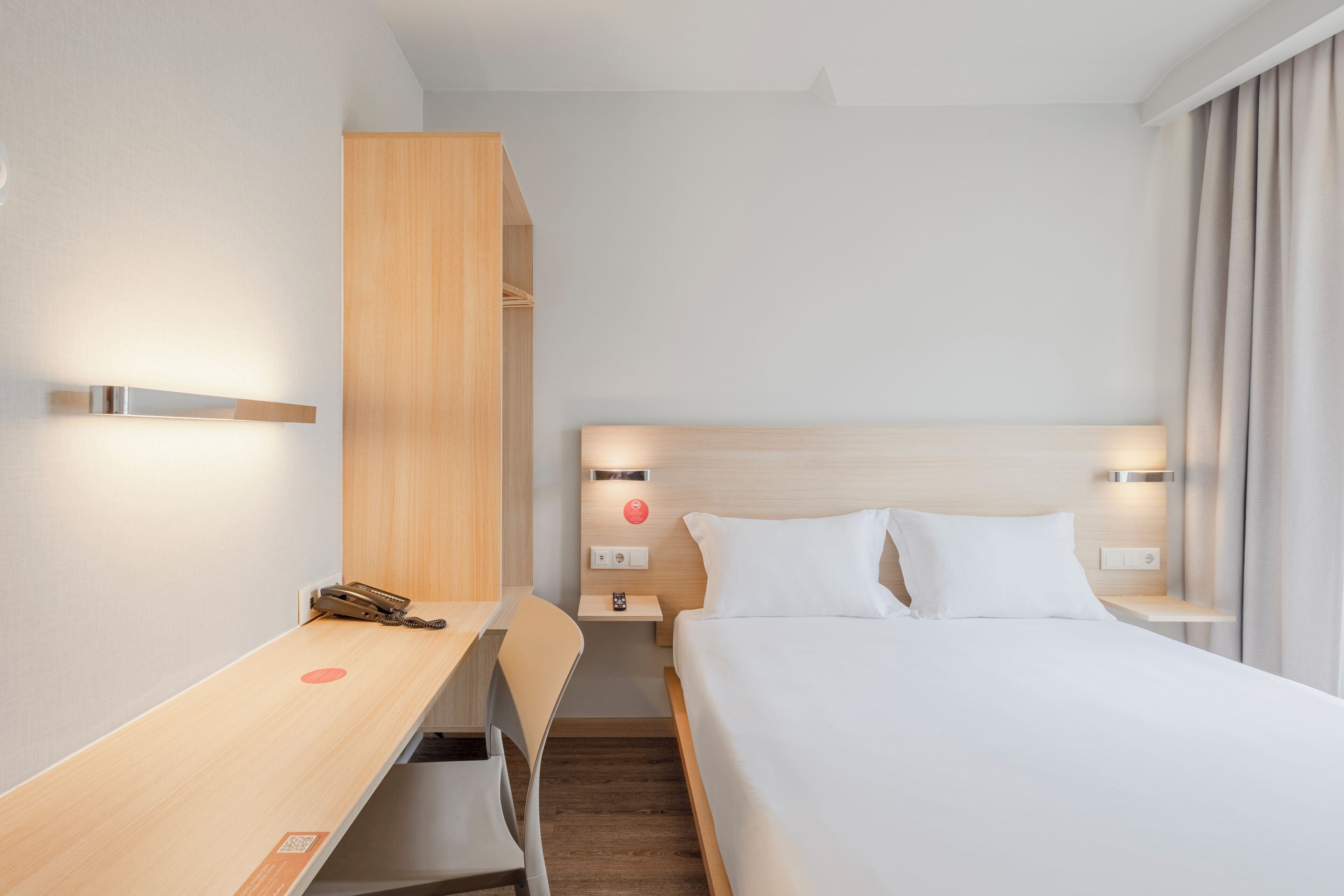 A hotel room with a bed, a desk, and a chair. There is a nightstand with a lamp and a phone.