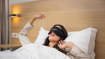 A person lying in bed with a black blindfold, white bedding, and a wooden headboard.