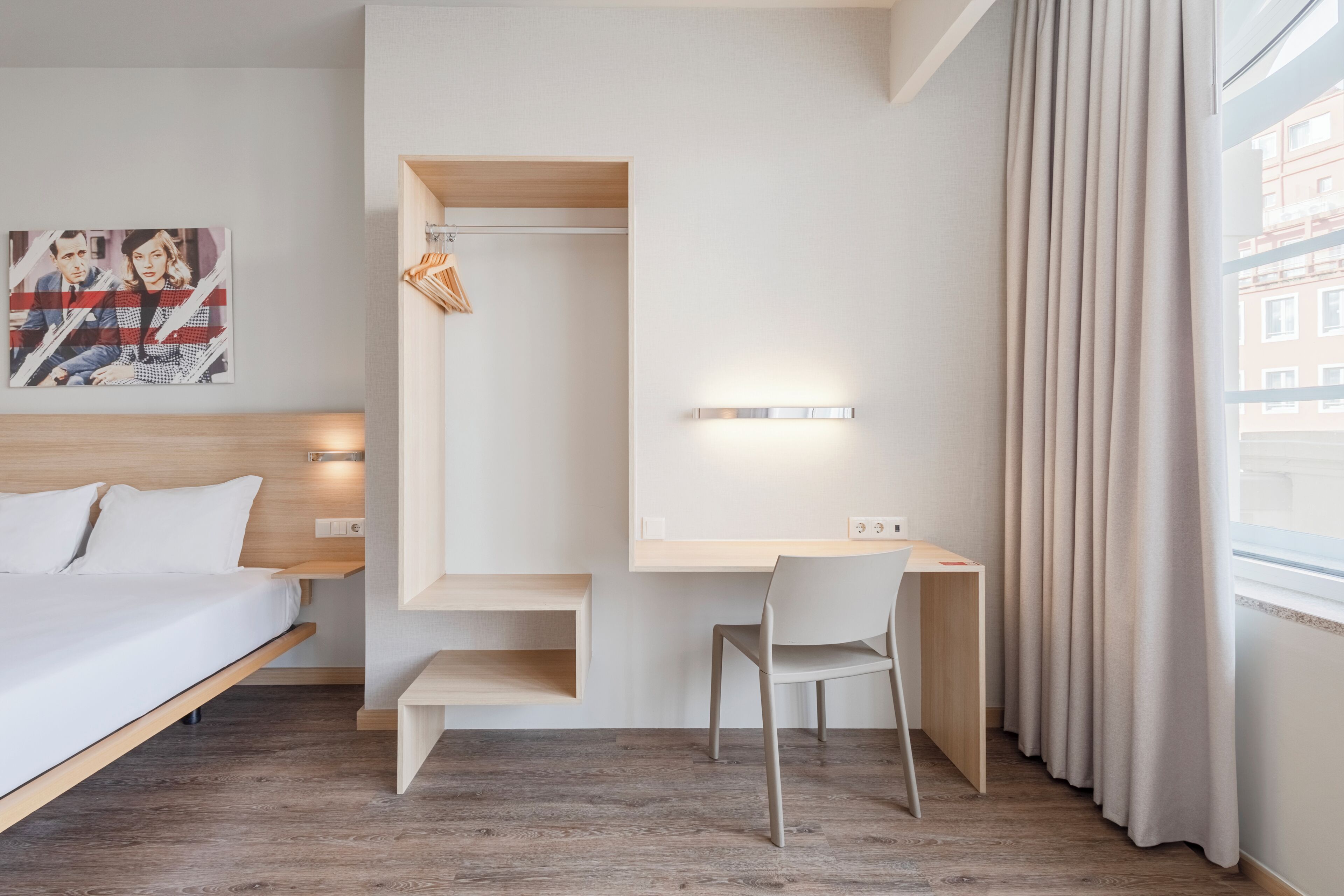 A modern hotel room with a bed, desk, chair, and a painting on the wall.