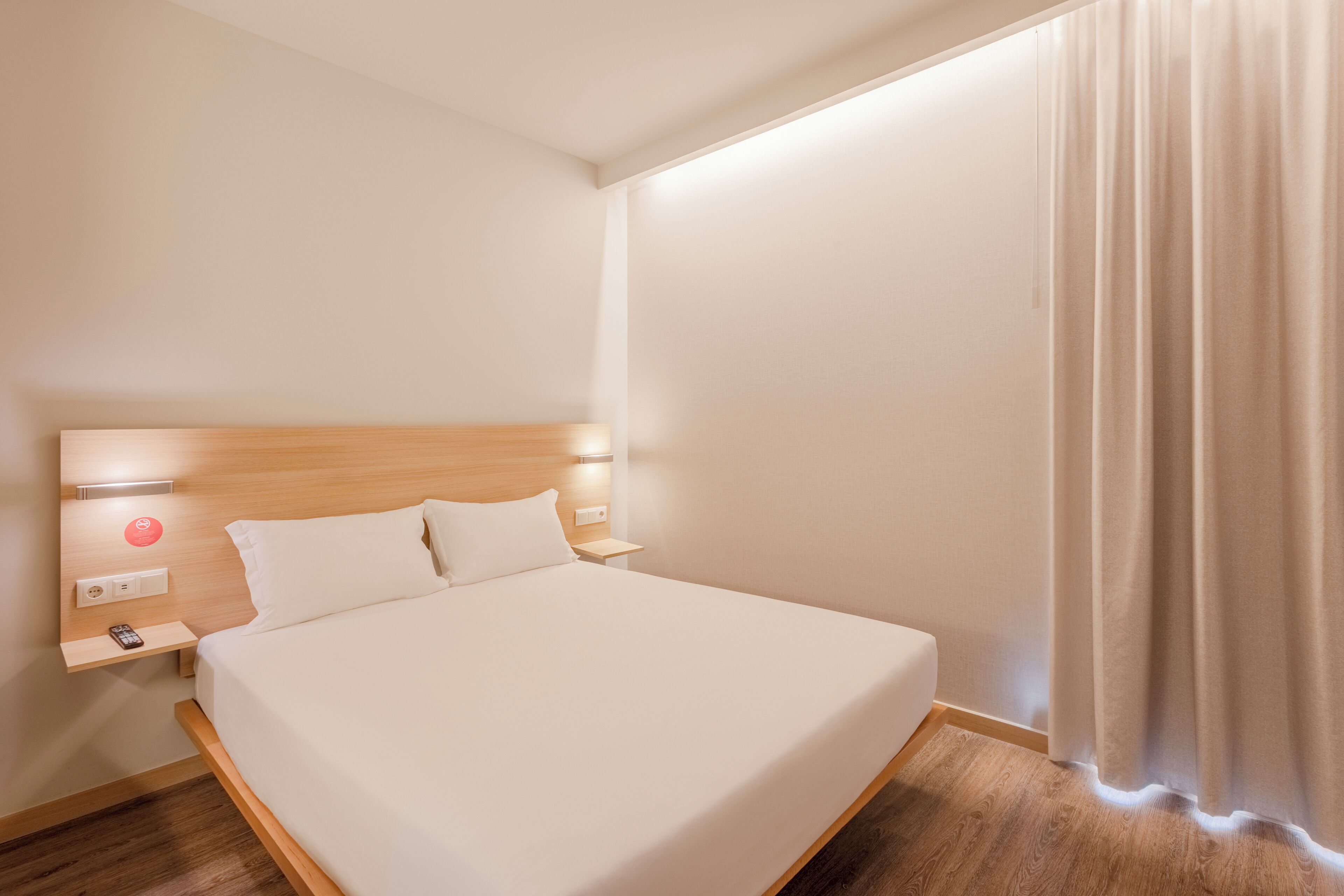 A neatly made bed with a wooden headboard, a bedside table with a phone and a red button, and a curtain on the window.