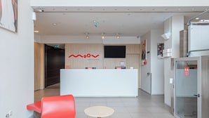 A modern hotel lobby with a reception desk, red seating, and a glass elevator. - Moov Hotel Porto Centro (Porto)